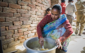 A woman in a sari crouches on the floor stirring a large pot of food