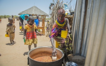 A woman in colourful clothing sitting outside is serving beans from a large pot while children wait beside it