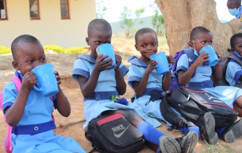 Five children sit in a line on the ground drinking from blue mugs