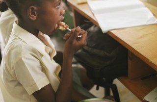 A child sitting at a desk, eating from a bowl of rice on her lap