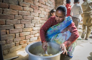 A woman in a sari crouches on the floor stirring a large pot of food