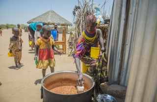 A woman in colourful clothing sitting outside is serving beans from a large pot while children wait beside it
