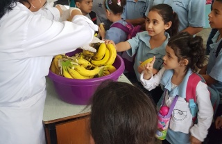 A group of adults stand behind a table which has large bowls full of bananas on it. A crowd of children stand in front of the table.