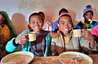 Two boys sit at a desk with bowls in front of them holding mugs up to the camera