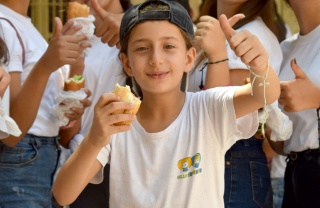 A child in a white t-shirt and backwards cap holds up a sandwich and gives the camera a thumbs up