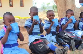 Five children sit in a line on the ground drinking from blue mugs