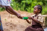 A woman bends down to hand a small child sitting on an overturned basket a bowl as they smile at each other