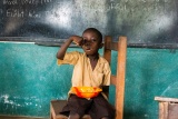 A child sits on a chair in front of a blackboard with a bowl on their lap, putting a spoonful of food into their mouth