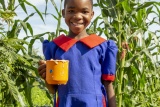 A child holding a mug standing in front of tall green plants and smiling at the camera