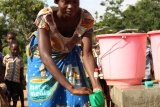 A woman wearing a Mary's Meals apron and hat leans over a bucket, washing a mug