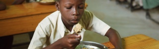 A boy sitting at a desk with a bowl of rice on it, blowing on a spoonful