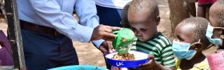 A child looks down at a bowl as beans are being poured into it by an adult