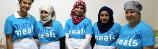 A group of five women smiling at the camera wearing Mary's Meals t-shirts and white aprons