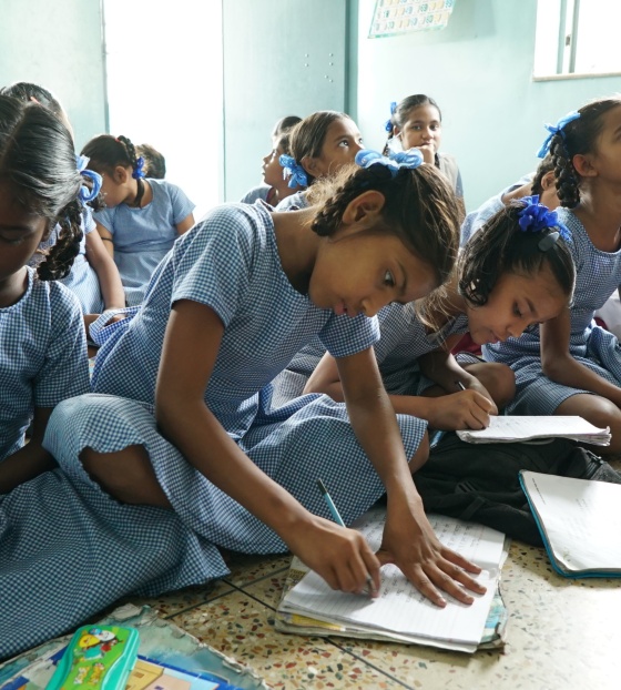 A room full of girls in school uniform sit on the floor, leaning over books they are writing in