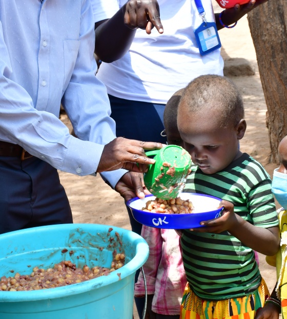 A child looks down at a bowl as beans are being poured into it by an adult