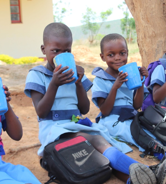 Five children sit in a line on the ground drinking from blue mugs