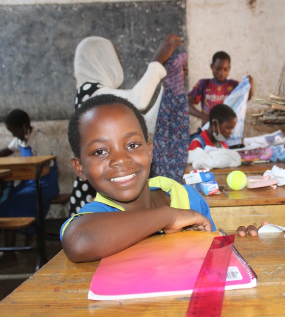 Young girl in class at her desk with a pink booklet in front of her smiling at the camera