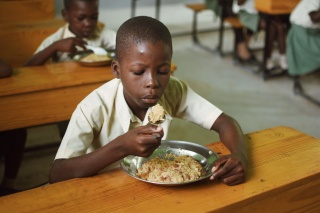 A boy sitting at a desk with a bowl of rice on it, blowing on a spoonful