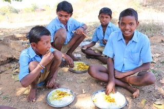 Four boys in school uniform sit on the ground with bowls of rice and curry in front of them