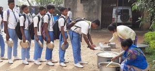 A group of boys wearing school uniform stand in a line waiting to be served food out of a large pot
