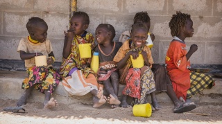 A group of six children sit on the ground holding yellow mugs and laughing