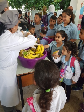 A group of adults stand behind a table which has large bowls full of bananas on it. A crowd of children stand in front of the table.