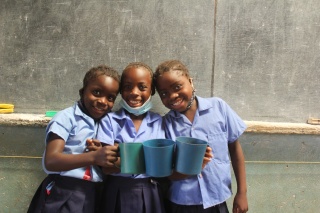 Three children wearing school uniform stand in front of a blackboard holding up blue mugs and smiling at the camera