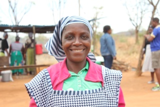 A woman wearing a pinafore and head scarf standing outside smiles at the camera