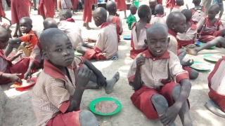 Children sitting on the dry ground covered in dust and eating bowls of porridge